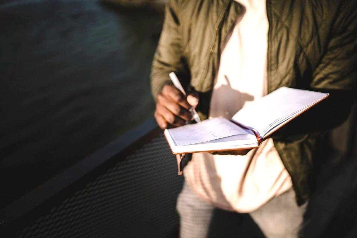 Persona de pie escribiendo a mano en un cuaderno, con la luz de frente y el rostro fuera del encuadre