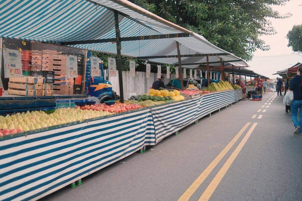 Puesto de fruta al aire libre: una mesa larga cubierta con un mantel a rayas y, encima, cajones de fruta ordenados a lo largo de la calle